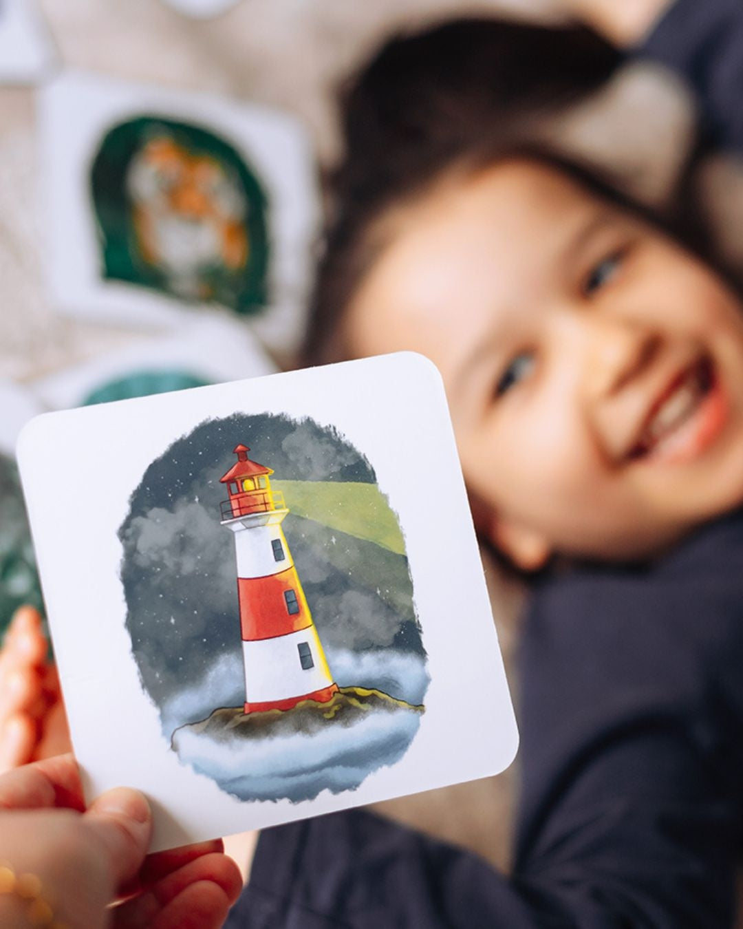 smiling child holding phonics and storytelling card showing a lighthouse illustration.