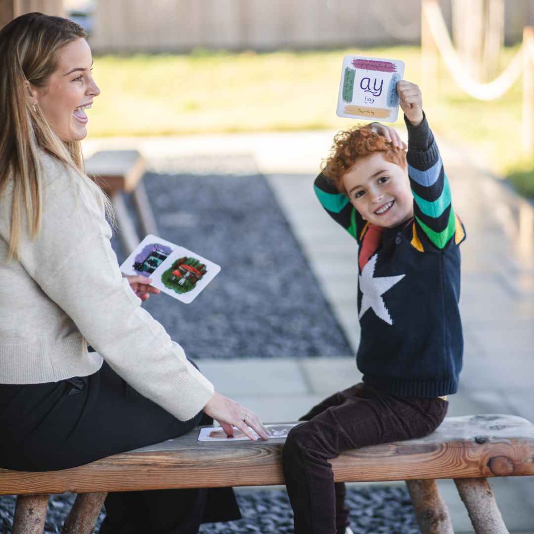 Child holding up phonics card while sat on a bench with his mother.