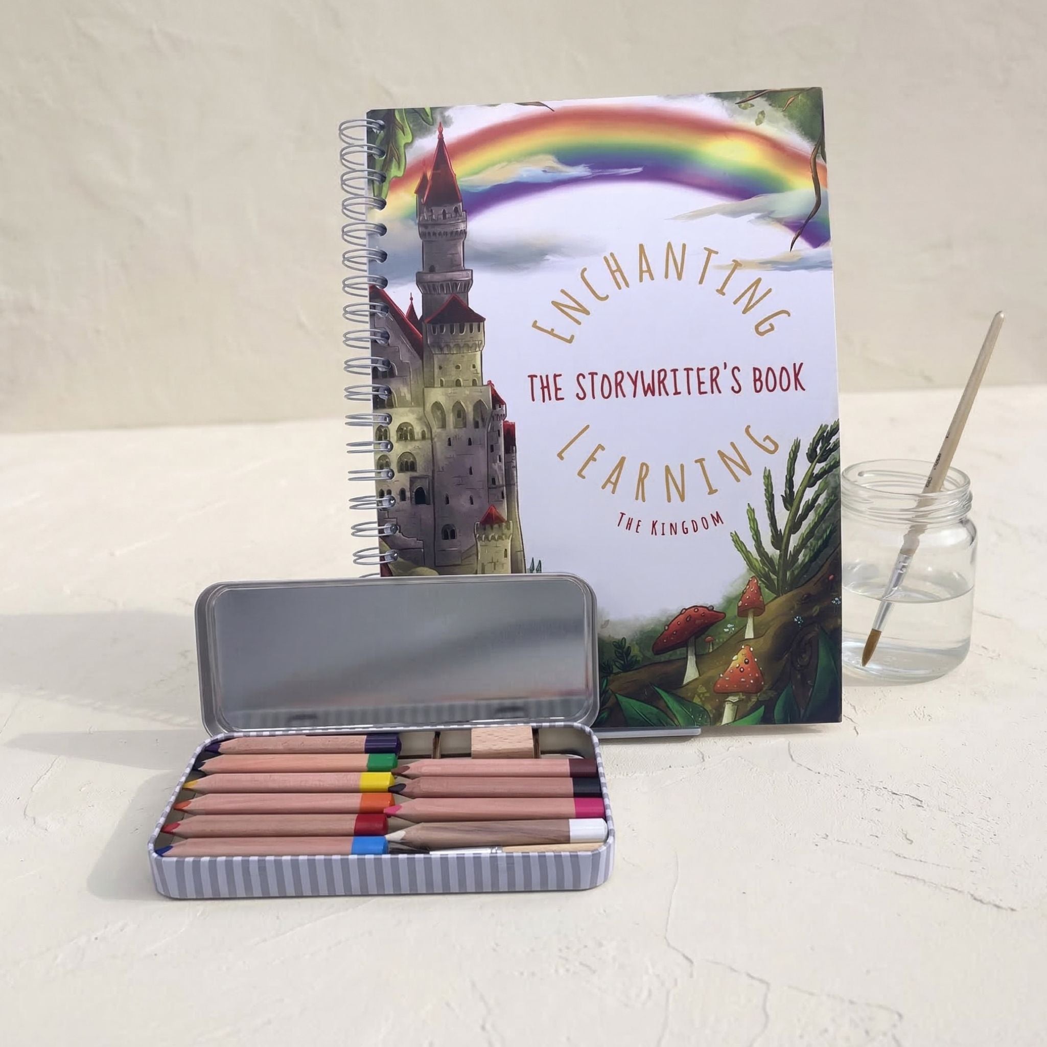 Storywriter's book with a castle illustration, colored pencils in a tin, and a jar with a paintbrush on a light surface.