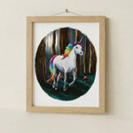 Framed artwork of a unicorn with a rainbow mane and tail in a forest setting.